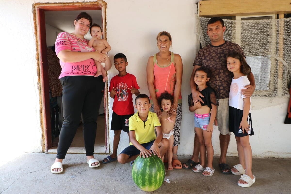 De pie tres adultos (dos mujeres y un hombre). Una de las mujeres carga en brazos a un bebé, y debajo de ellos hay un bebé más, dos niñas y dos niños.