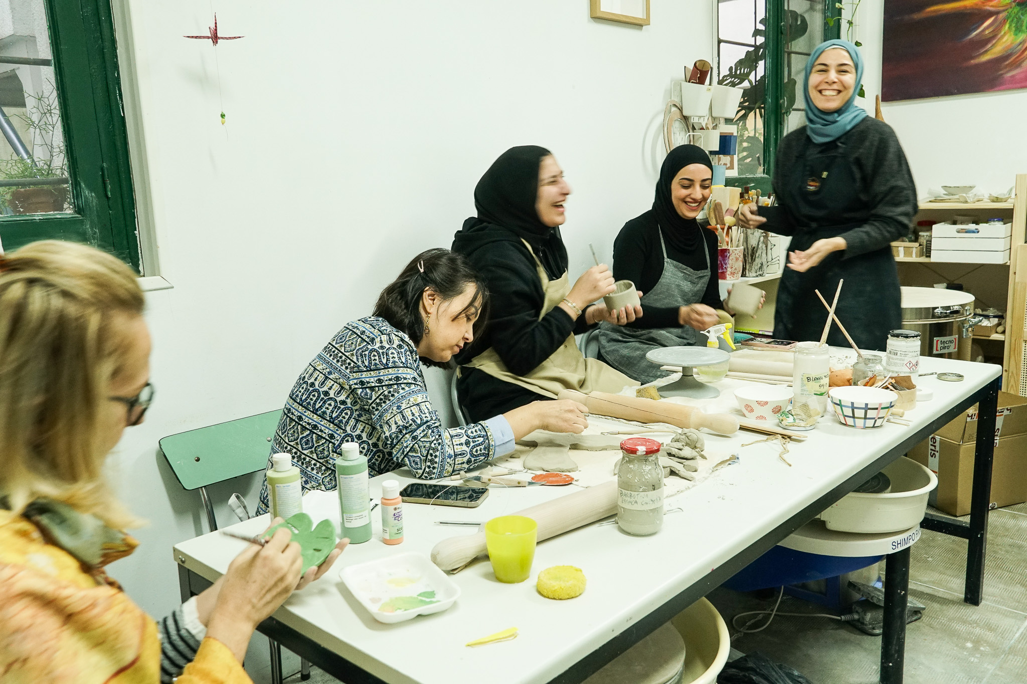 Cinco mujeres trabajan y r&iacute;en en torno a una mesa con materiales de cer&aacute;mica.