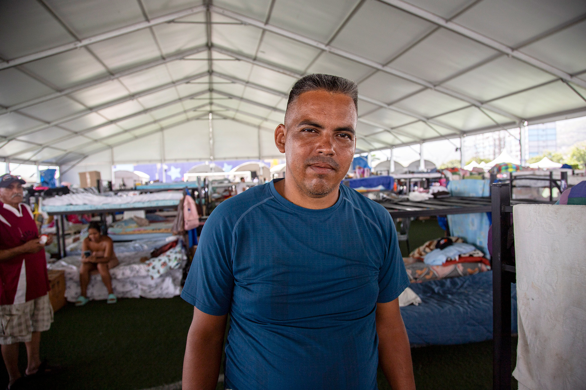 Un hombre que viste camiseta azul, está de pie en un albergue temporal. Detrás de él se observan decenas de literas