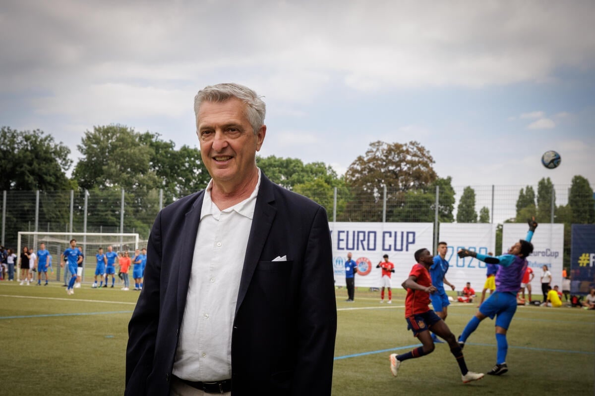 Un hombre cano en una cancha de f&uacute;tbol sonr&iacute;e a la c&aacute;mara. Detr&aacute;s suyo, dos equipos jugando. 