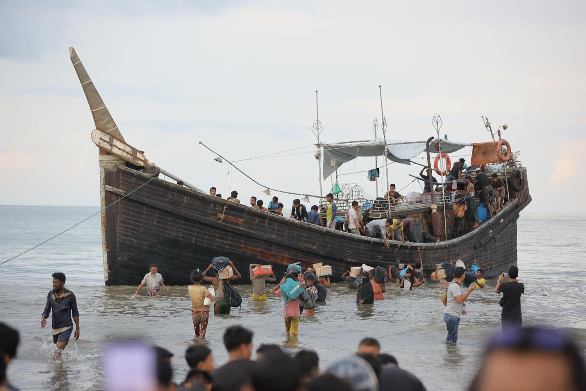 Un grupo de personas con sus pertenencias se encuentra en aguas poco profundas junto a un barco de madera que transporta decenas de personas