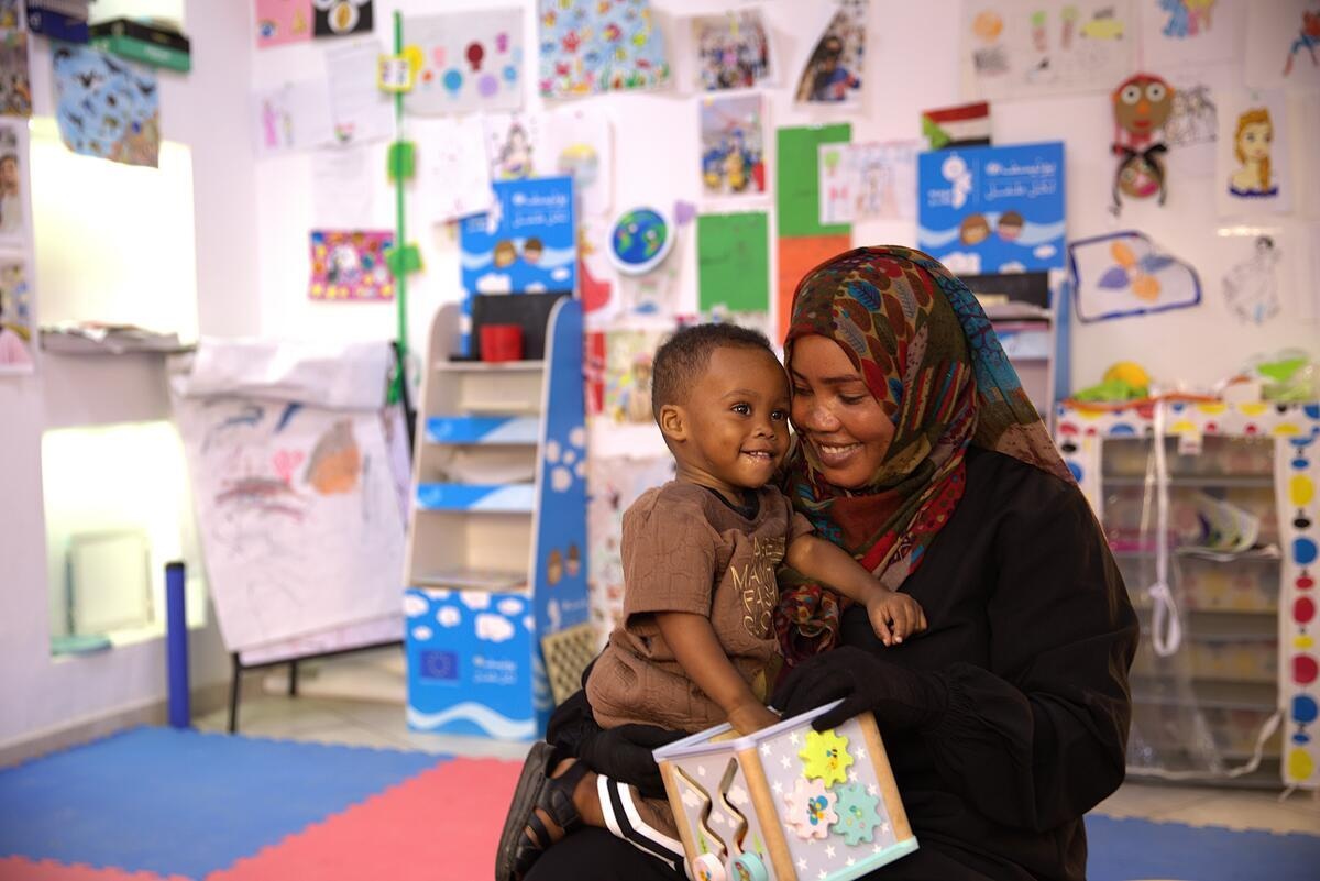 En medio de un aula, una mujer sudanesa sostiene a su hijo en brazos; ambos sonríen. En las paredes hay ilustraciones que parecen haber sido elaboradas por niñas y niños.