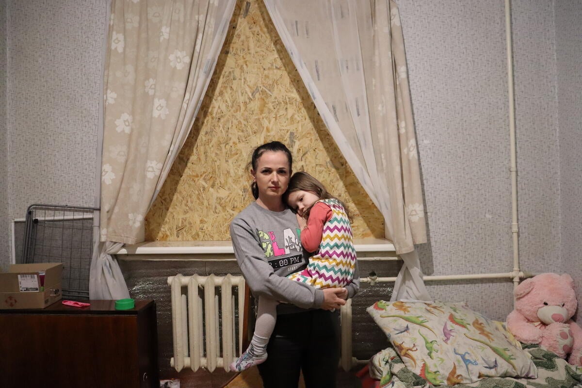 Una madre carga a su hija pequeña en brazos frente a una ventana tapada con madera en la habitación de su hija