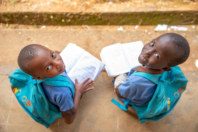 Dos niñas de 7 y 8 años, procedentes de la República Centroafricana, sonríen a la cámara desde donde están sentadas al aire libre, con mochilas a juego en las que se lee 'La educación no puede esperar' y sosteniendo libros abiertos