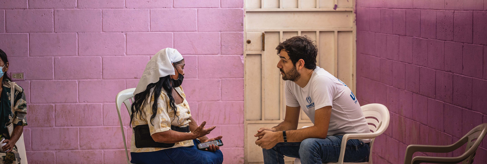 Un trabajador de ACNUR con camiseta blanca y logos azules, platica con una mujer que usa un pañuelo blanco en la cabeza