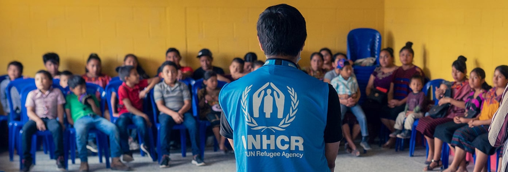 Foto de portada de página: un hombre de espaldas frente a un grupo de personas, muestra el chaleco azul con visibilidad de ACNUR