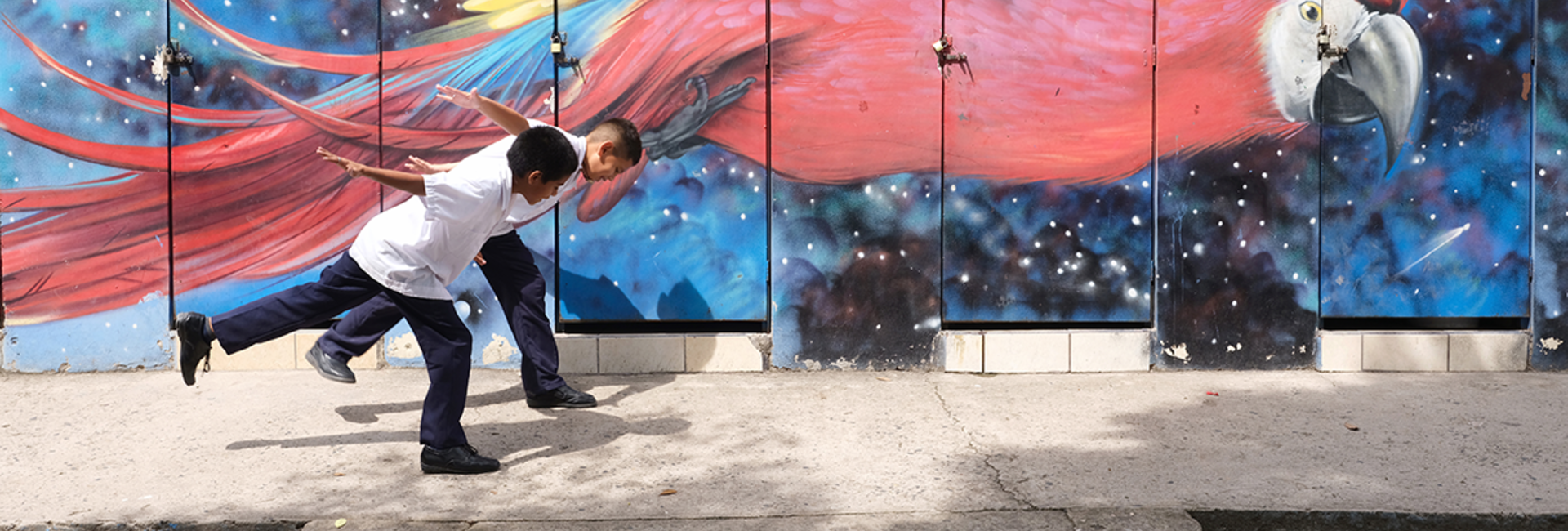 Dos niños juegan sobre la acera; detrás suyo, un mural en el que figura una ave de color rojo.