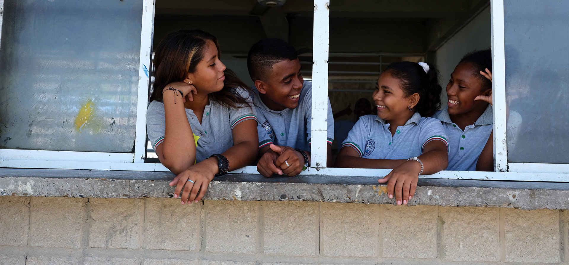 Cuatro adolescentes (tres niñas y un niño) que visten camiseta de uniforme escolar gris, se asoman por la ventana de un salón de clases