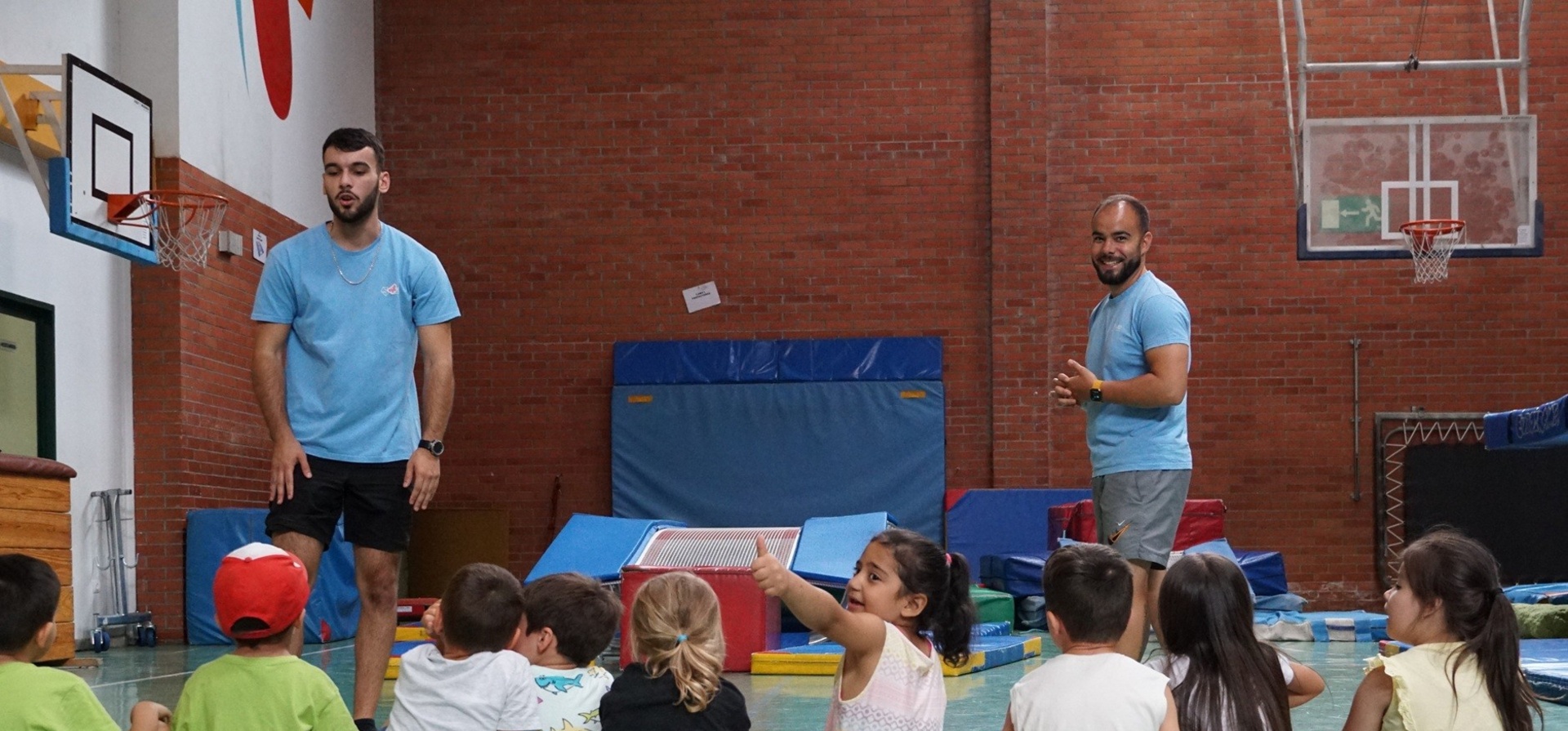 Un grupo de niñas y niños sentados en un polideportivo frente a dos monitores.