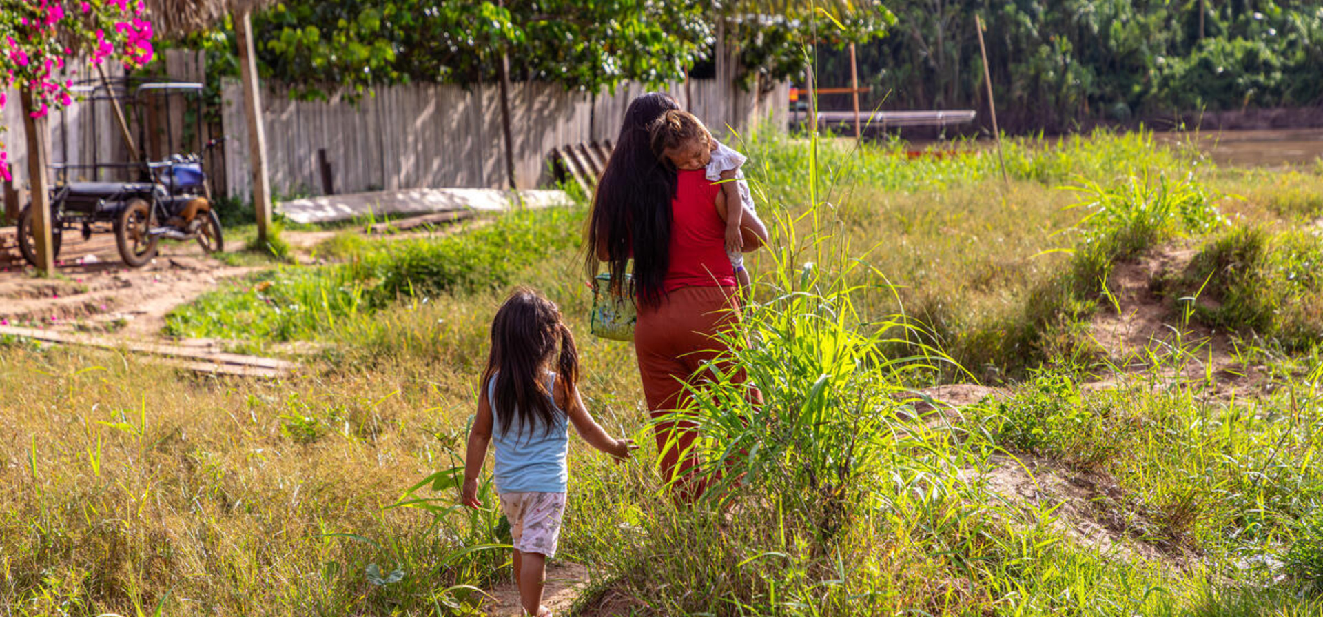 Una mujer camina de espaldas cargando en sus brazos a una niña, mientras otra niña las sigue de cerca.