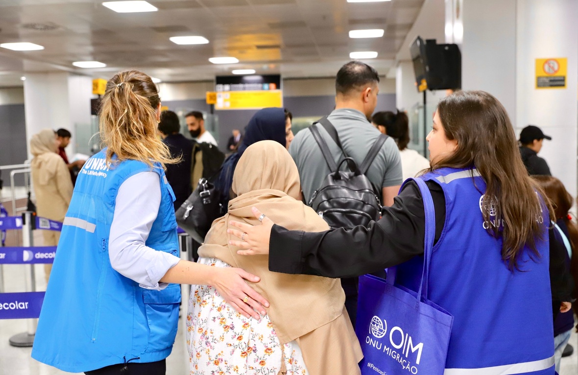 Dos mujeres que llevan puestos chalecos azules acompañan a una mujer que lleva un velo en su cabeza en un aeropuerto.