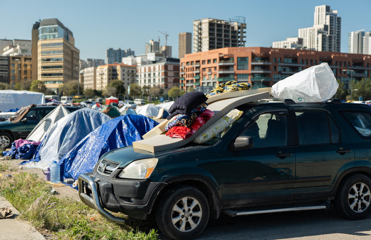 Tiendas de campaña y un vehículo con colchones y almohadas en un lote de estacionamiento cerca de algunos edificios.