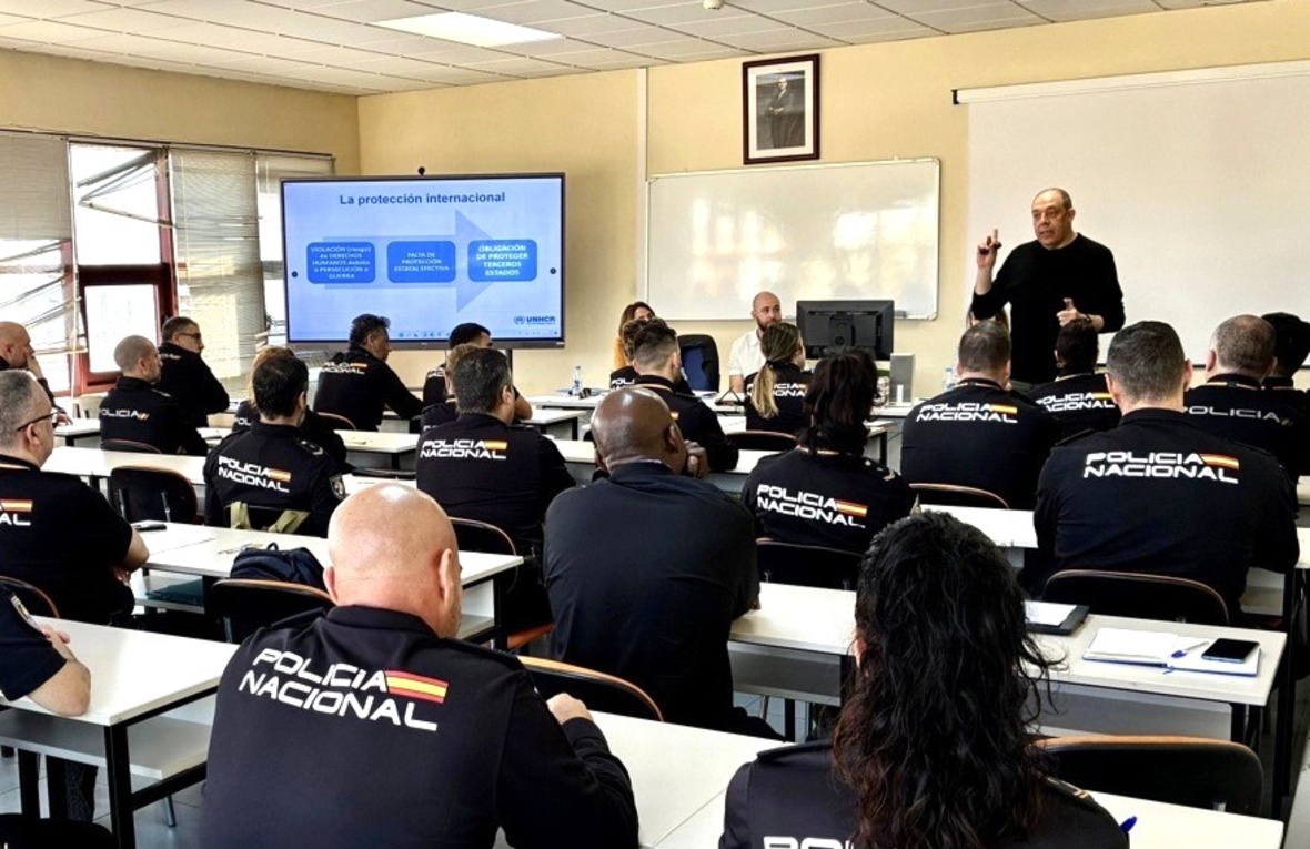 Un aula llena de agentes con uniformes de policía escucha a un hombre vestido con jersey oscuro.