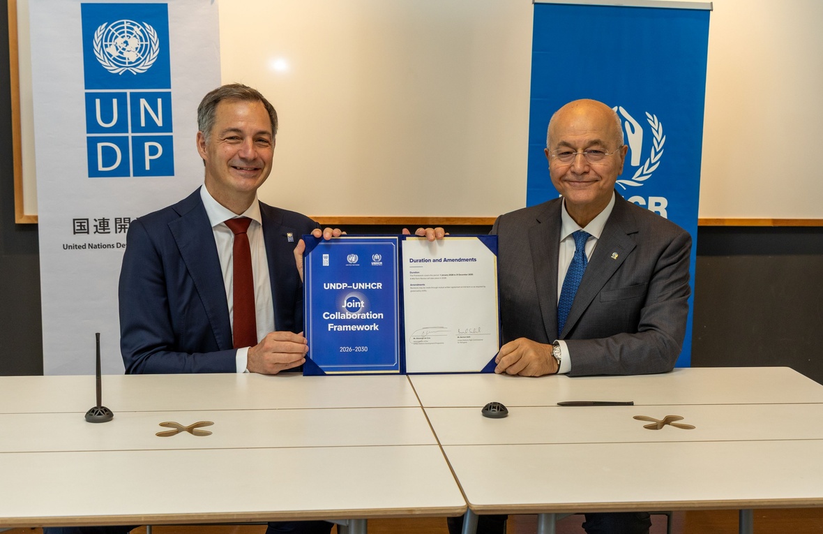 Dos hombres sonrientes que están sentados a una mesa sostienen un documento firmado en el que se lee “Acuerdo de colaboración conjunta entre el PNUD y ACNUR: 2026-2030”. Detrás de ellos se ven pancartas del PNUD y de ACNUR