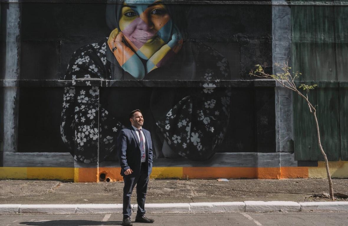 Un hombre vestido con traje y corbata de pie en una calle frente a un muro negro con el rostro de una mujer en colores
