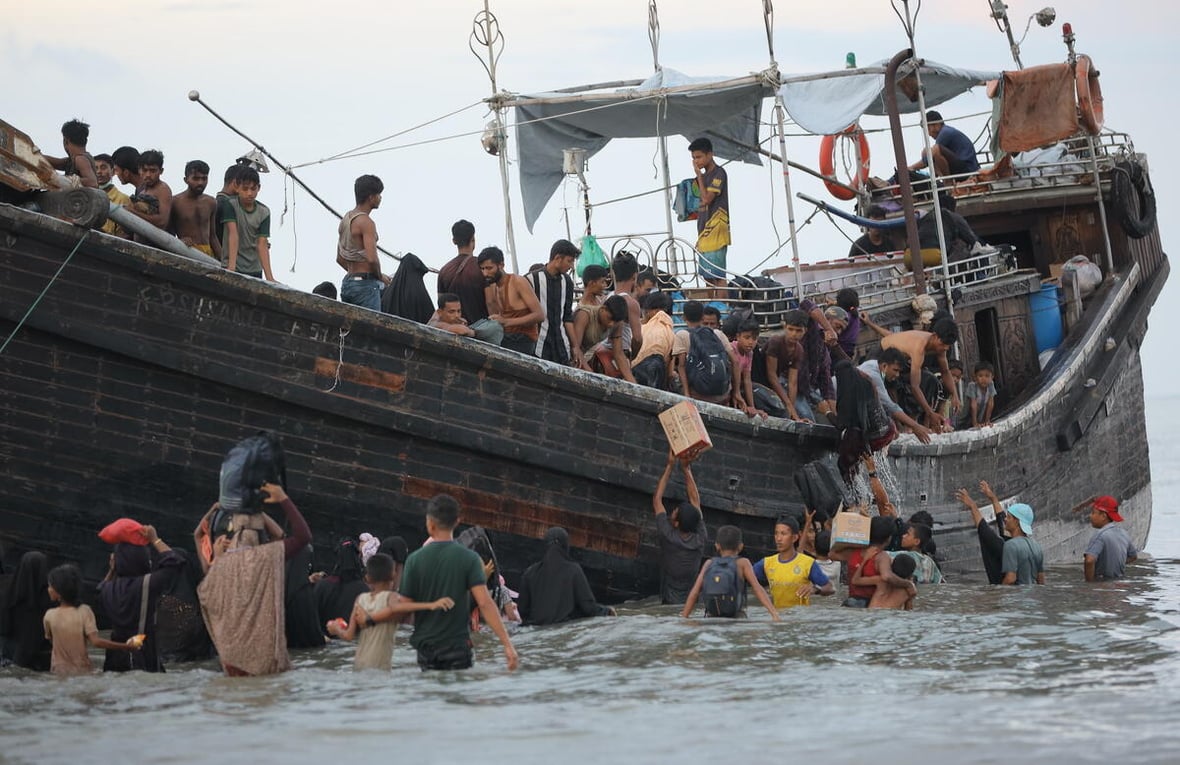 Decenas de personas se ven a bordo de una destartalada embaracación y de pie en las aguas bajas que la rodean, algunos ayudando a otros a bajarse del barco, otros más descargando bultos y cajas.
