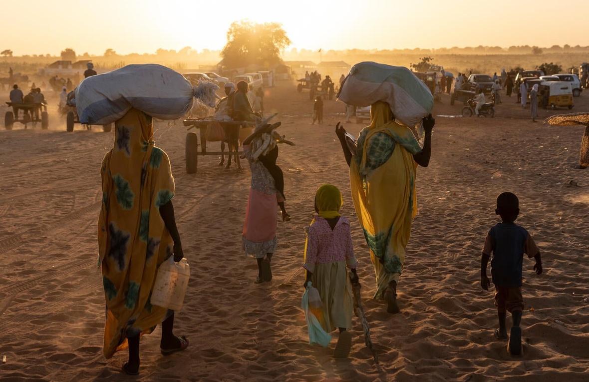 Personas refugiadas sudanesas y sus hijos caminan hacia el sol, llevando sus pertenencias sobre la cabeza o en carretas