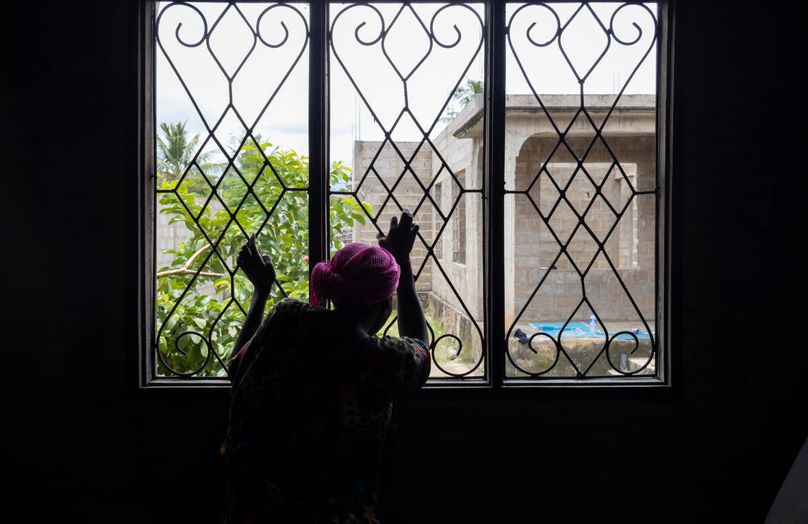 Una mujer de espaldas a la cámara mira por una ventana con las manos en los barrotes de seguridad.