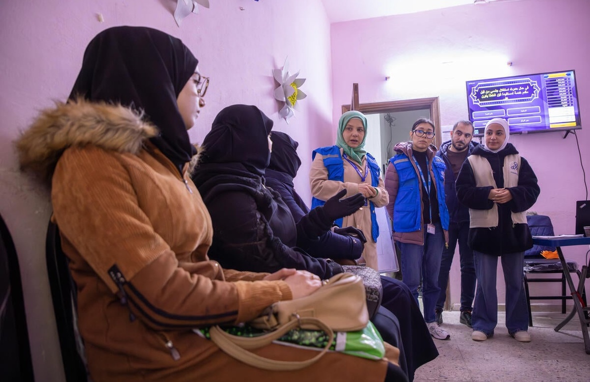 Cuatro personas de pie con uniforme de ACNUR se dirigen a 3 mujeres con la cabeza cubierta que están sentadas en sillas 