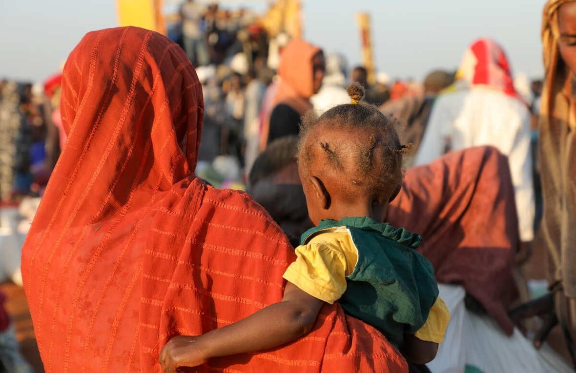Una mujer sudanesa lleva en brazos a su bebé; se les va de espaldas. Delante suyo, un numeroso grupo de personas.