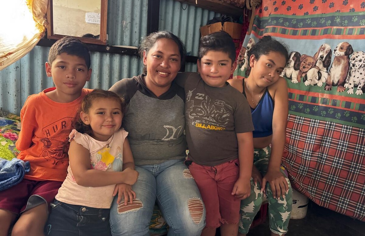 Una mujer sentada sobre una cama con sus cuatro hijos (dos niñas y dos niños). La familia sonríe a la cámara.