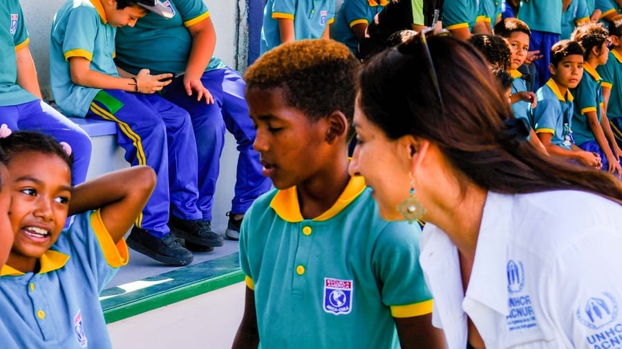 Tres niños con uniforme escolar azul con amarillo conversan con una trabajadora de ACNUR que viste camisa blanca con logos azules de ACNUR