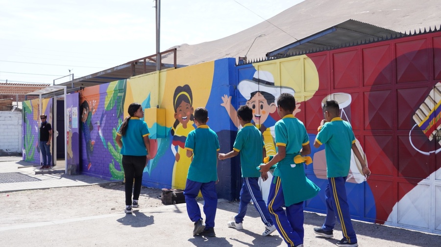 Cuatro niños y una niña caminan al lado de un mural colorido en una escuela