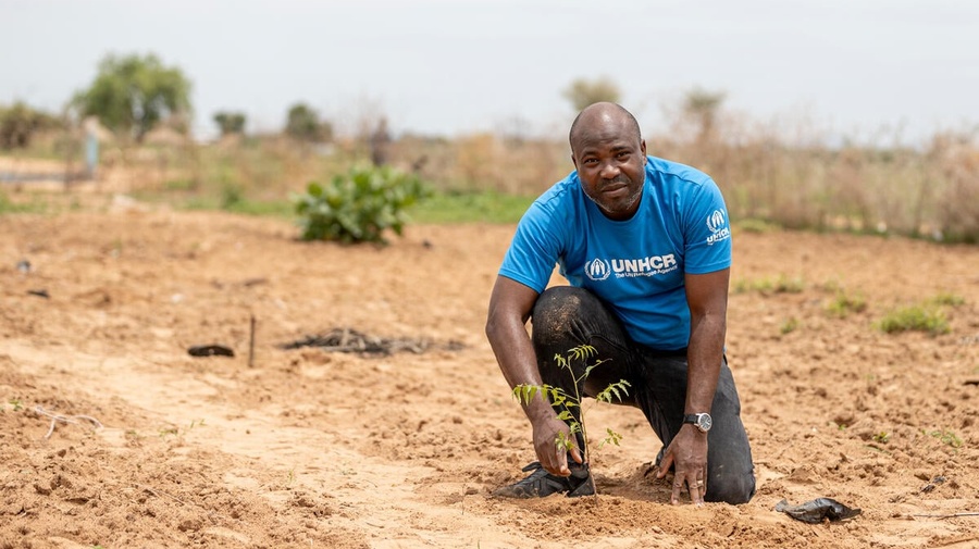 Un hombre en Camerún ve fijamente a la cámara. Lleva puesta una camiseta con el logo de ACNUR y están hincado en una tierra de cultivo.