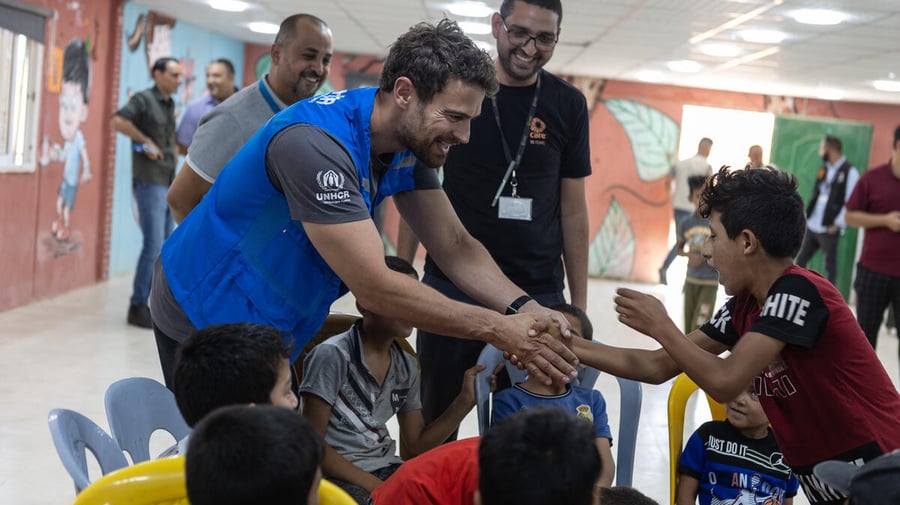 Un hombre joven que lleva puesto un chaleco con el logotipo de ACNUR salud a un grupo de niños en un campamento de refugiados en Jordania