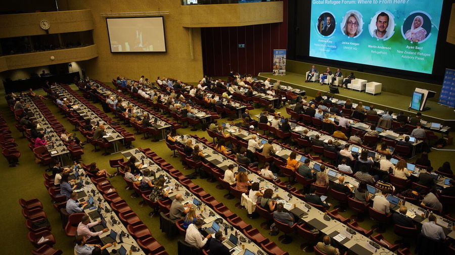 Imagen de un auditorio repleto de personas; al frente, un panel y una pantalla en la que se está proyectando una presentación