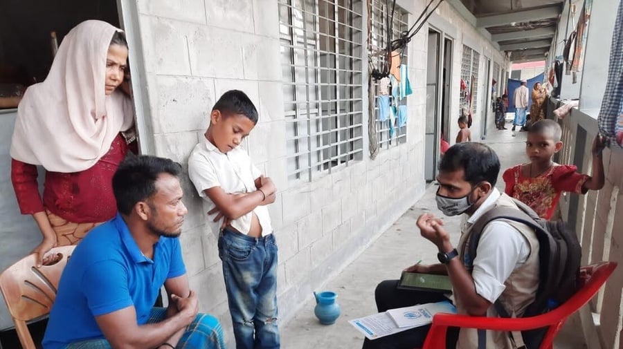 Un trabajador humanitario charla con una familia: el padre está sentado en una silla en la puerta del hogar; detrás suyo, la madre, quien lleva puesto un velo.
