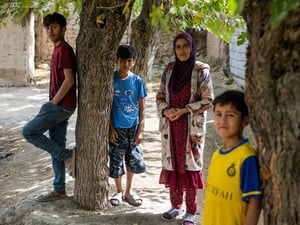 Una mujer con la cabeza cubierta con un pañuelo morado entre unos árboles con tres niños de distintas edades que visten camisetas color vino, azul y amarillo