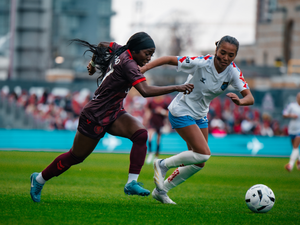 Dos mujeres jóvenes vestidas con uniformes de fútbol de equipos diferentes, corren detrás de un balón en una cancha con pasto verde