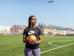 Una joven sostiene un balón de fútbol en una cancha de fútbol con hileras de casas al fondo