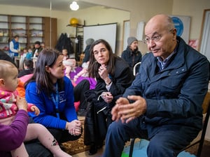 Un hombre habla con una mujer que sostiene a una bebé en su regazo, sentados en una sala común con otras personas al fondo