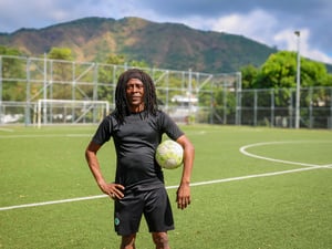 Un hombre de piel oscura y cabello largo oscuro vestido de negro, posa para la foto en una cancha de fútbol con pasto verde mientras sostiene con su brazo izquierdo un balón de fútbol