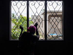 Una mujer de espaldas a la cámara mira por una ventana con las manos en los barrotes de seguridad.