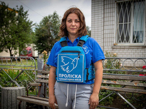 Una mujer que viste camiseta azul, sostiene al frente de ella una mochila azul con letras blancas, mientras se encuentra de pie frente a una banca de madera en un jardín