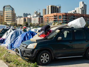 Tiendas de campaña y un vehículo con colchones y almohadas en un lote de estacionamiento cerca de algunos edificios.
