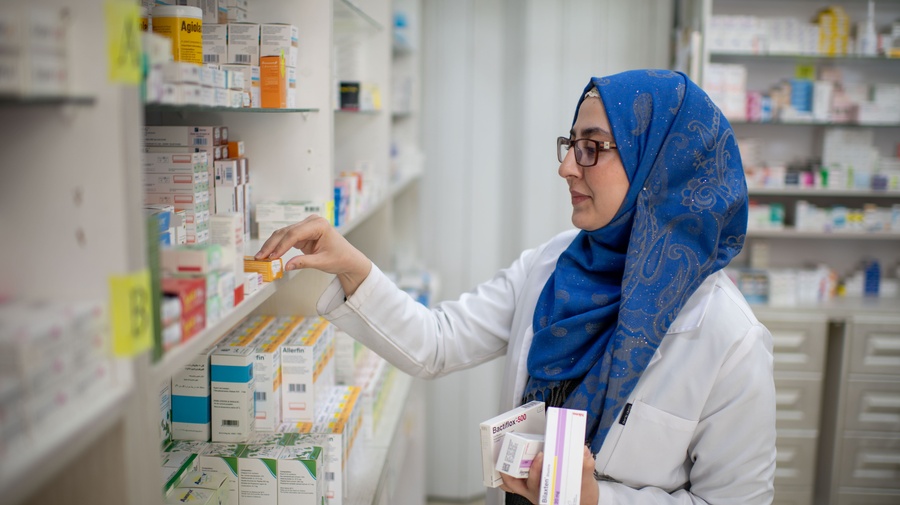 Una mujer joven, que usa anteojos y lleva puesto un hiyab azul y una bata de laboratorio, acomoda medicamentos en un estante