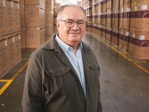 Un hombre que viste chaqueta café y camisa azul, posa para la foto en un pasillo de una bodega frente a cajas de cartón apiladas