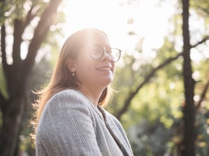 La luz del sol brilla a través de los árboles sobre una mujer que lleva lentes y un blazer gris