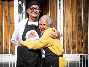 Un señor y una señora mayores que lleva puestos unos delantales de cocina posan para la cámara abrazándose y sonriendo.