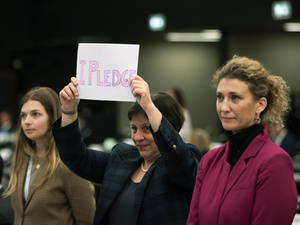 Tres mujeres forman una hilera. Las que están a los extremos son rubias; la que está en el centro, de cabello corto y oscuro, sostiene frente a sí un cartel que dice "Me comprometo".