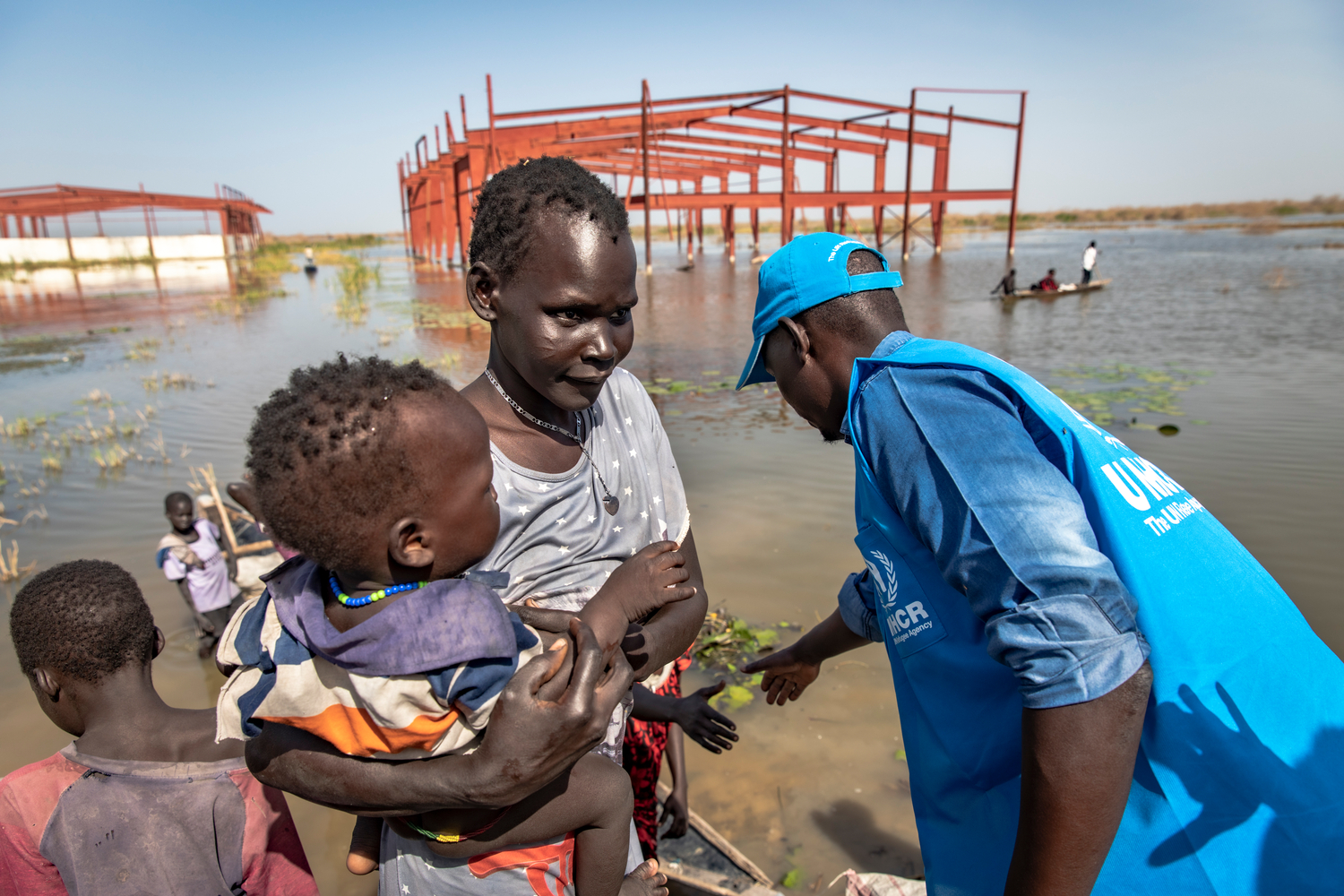 A man in a UNHCR vest help a mother and her children out of a canoe. Un hombre con chaleco de ACNUR ayuda a una madre y a sus hijos a salir de una canoa.