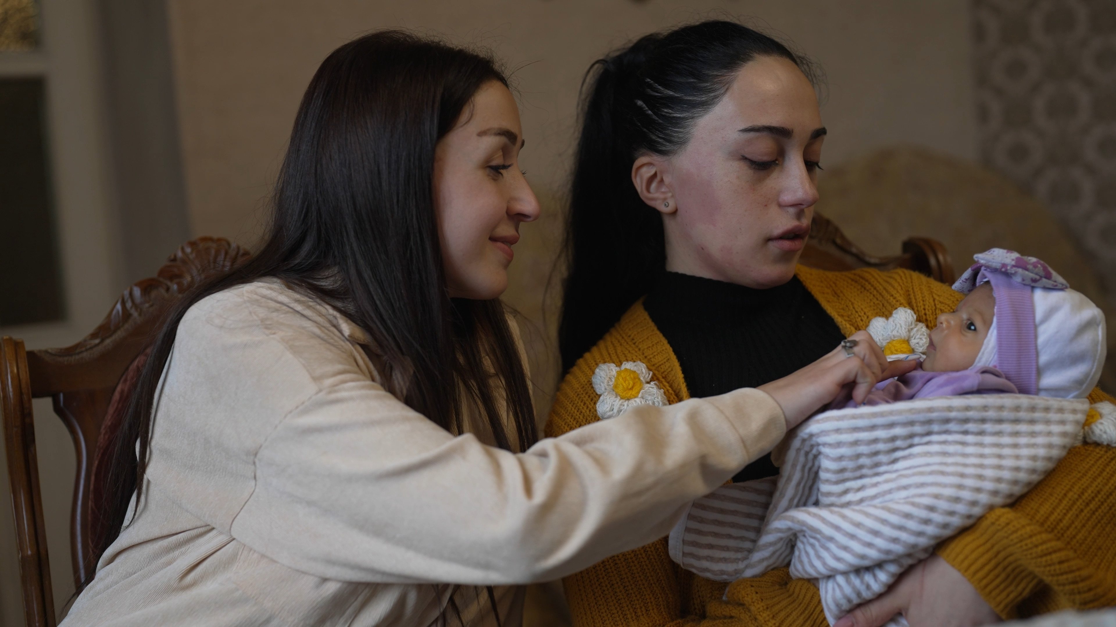 Dos mujeres observan a una bebé recién nacida, la cual es cargada por una de ellas que viste suéter amarillo.