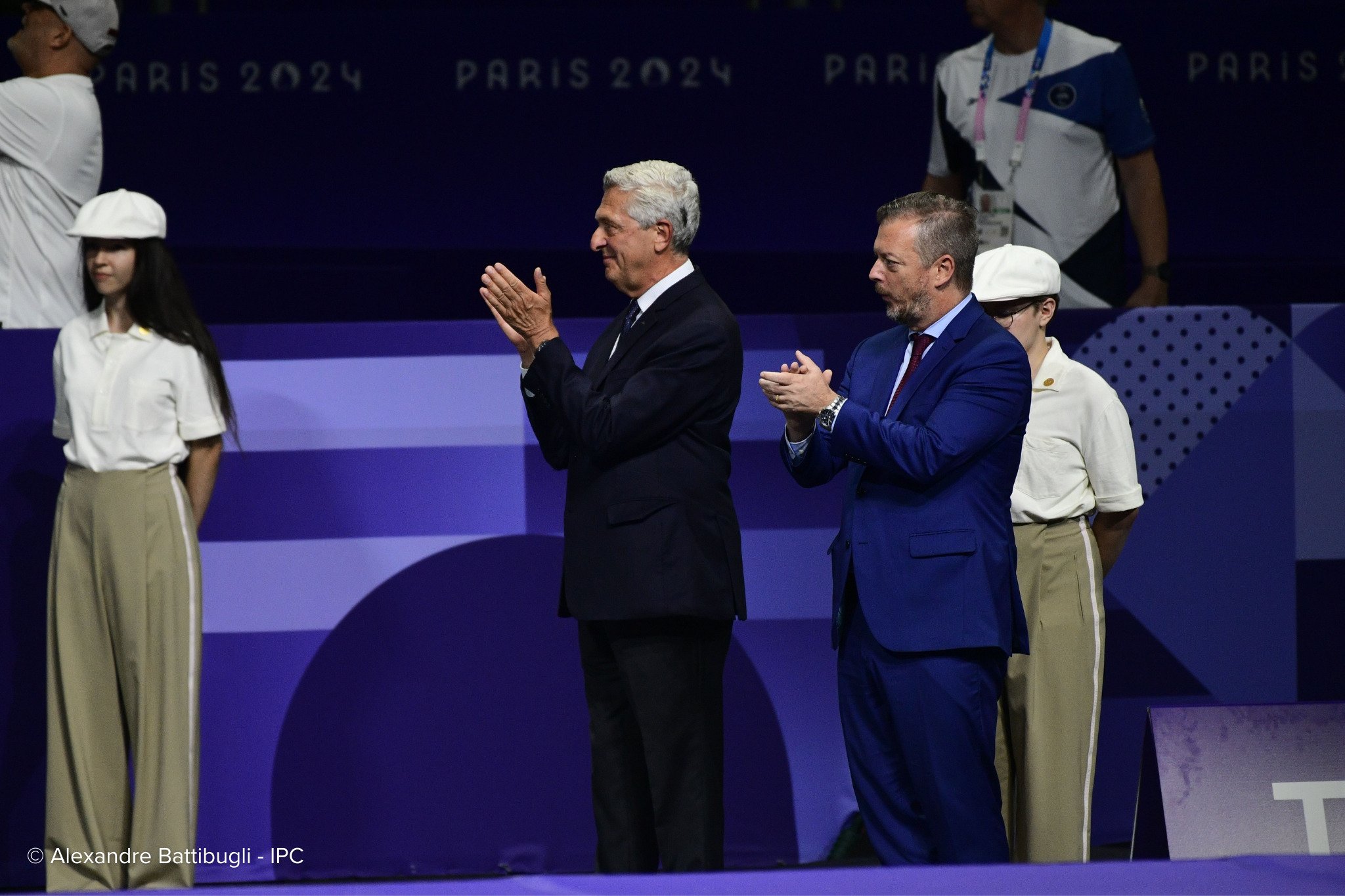 Dos hombres con traje aplauden en un lugar donde se exhiben los logotipos de los Juegos de París 2024