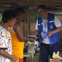 Un hombre con visibilidad de ACNUR en entrega de lámparas solares a dos hombres en una casa de madera.