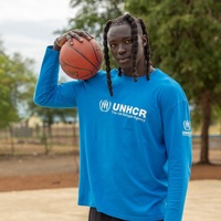 uN hombre con rastas en el cabello que viste una camiseta azul de ACNUR, sostiene una pelota de baloncesto color naranja en una cancha de baloncesto