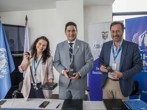 Una mujer y dos hombres durante la entrega de radios. Al fondo, banderas de la ONU y ACNUR